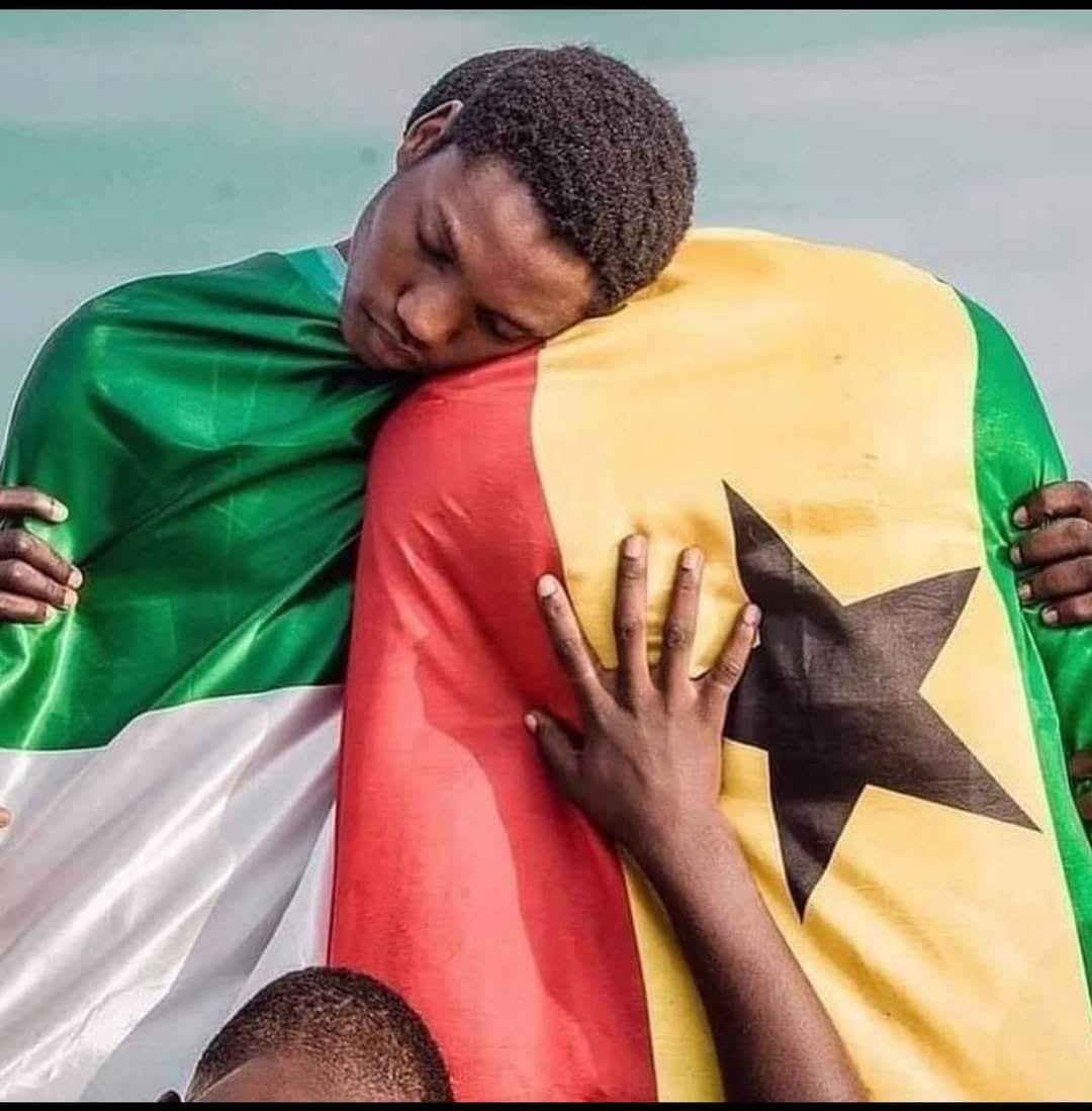 A young Ghanaian man wrapped in the national flag of Ghana, resting his head on a friend's shoulder