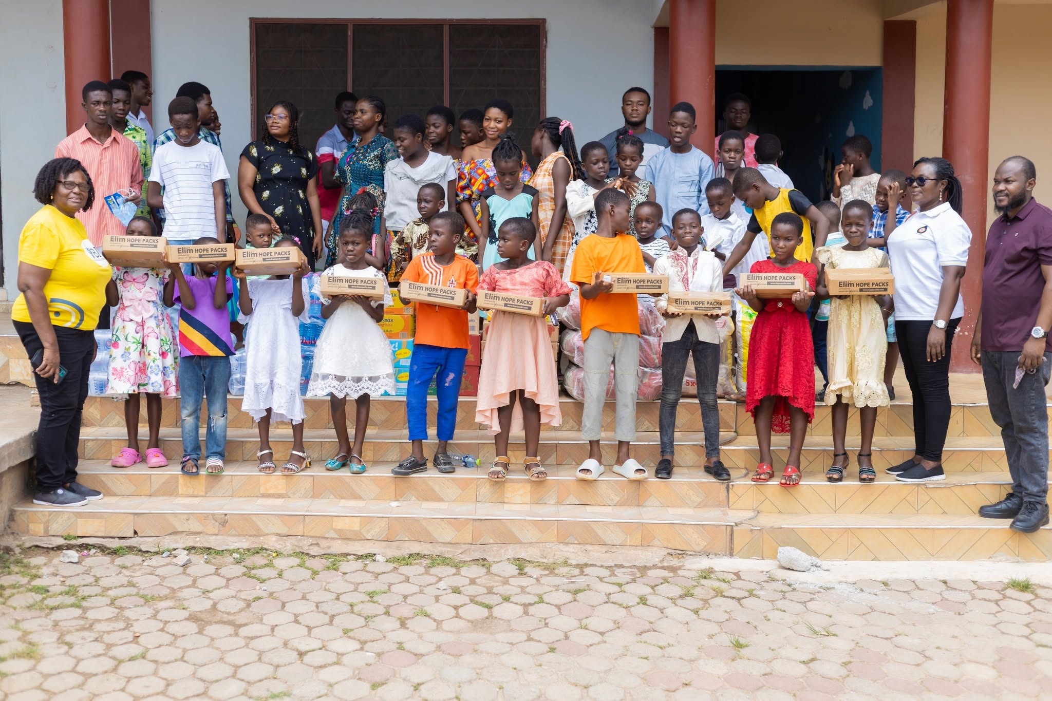 Children standing on school steps holding learning-kit boxes during an ACAI community outreach